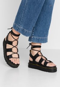 Svarte skinnsandaler med kryssede stropper og ankelband, med en tykk såle som har gule sømmer og åpne tær.