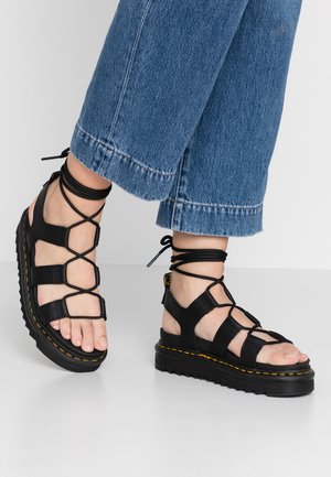 Black leather sandals with crisscross straps and ankle ties, featuring a thick sole with yellow stitching and open toes.
