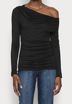 Person iført en sort langærmet top med rynkede detaljer, hvor den ene skulder er blotlagt, og blå jeans mod en ensfarvet baggrund.