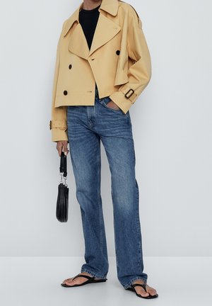 Veste beige courte à double boutonnage, haut noir, jean bleu à jambes droites, sac à main noir et sandales noires à semelle plate, main dans la poche.