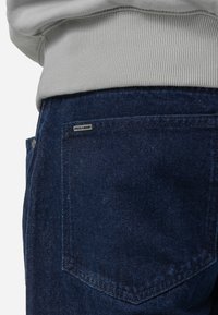 Sötétkék farmernadrág, amelyen egy jól látható hátsó zseb található, fekete 'PEGADOR' címkével. Az anyag textúrált megjelenésű, amely jól illik egy szürke pulóverhez.