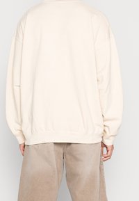 Osoba v přepychové krémové mikině a volných béžových kalhotách, otočená zády, na pozadí jednoduchého světlého pozadí.