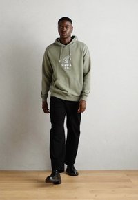 Sweat à capuche vert olive avec logo, poche kangourou à l'avant et cordon de serrage. Associé à un pantalon noir et des chaussures noires brillantes, sur un fond clair.
