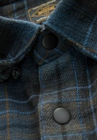 Detaliul gulerului cămășii din flanel în carouri negre și albastre, cu butoane negre și o textură țesută. Eticheta are inscripția "Durabil" într-un stil vintage.