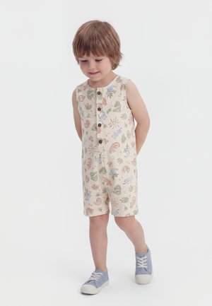 Malo dijete u bezbojnim kombinezonu bez rukava s printom životinja i biljaka, gleda dolje i hoda u plavim tenisicama.