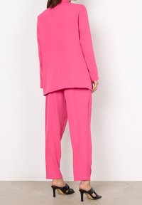 Różowy, luźny zestaw garniturowy składający się z długiego płaszcza i dopasowanych spodni o gładkiej teksturze. Całość dopełniają czarne sandały na obcasie.