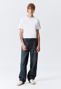 T-shirt branca de mangas curtas combinada com jeans de perna larga azul escuro. A modelo usa ténis brancos com detalhes em bege. Fundo é branco liso.