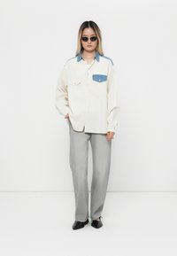 Junge Frau trägt ein übergroßes cremefarbenes Hemd mit blauen Denim-Details, graue gerade Jeans, schwarze Schuhe und Katzenaugen-Sonnenbrille, während sie vor einem weißen Hintergrund steht.