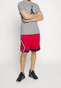 Šedé tričko s černým sportovním logem, doplněné červenými šortkami s bílými a černými detaily a černým pasem, nošeno s černými teniskami.