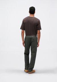 Άνδρας με την πλάτη στραμμένη, φορώντας καφέ μπλουζάκι, σκούρα πράσινα παντελόνια με ελαστική μέση και καφέ παπούτσια, σε απλό ανοιχτό φόντο.