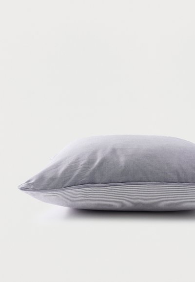 Γκρι υφασμάτινο μαξιλάρι με αδιάκριτο πάνω μέρος και ριγέ κάτω μέρος, που ακουμπά σε λευκή επιφάνεια μπροστά σε απλό φόντο.