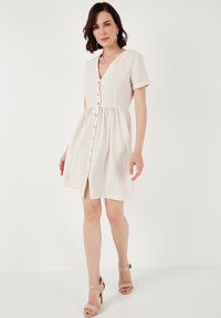 Light beige short-sleeve dress with a button-down front, v-neckline, and gathered waist, featuring a flowing skirt and rolled cuffs.