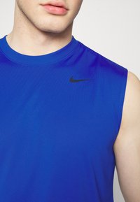 Canottiera atletica blu senza maniche realizzata in un tessuto liscio, con un logo Nike nero sul lato sinistro del petto. Collo rotondo semplice, design aderente.