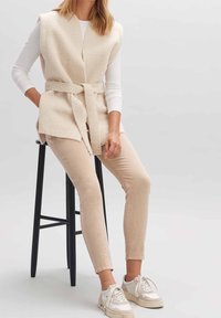 Femme assise sur un tabouret noir portant un haut blanc à manches longues, un gilet polaire beige ceinturé, un pantalon beige et des baskets blanches.