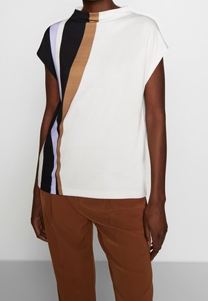 Person trägt ein ärmelloses weißes Oberteil mit vertikalen schwarzen, braunen und lavendelfarbenen Streifen auf einer Seite, kombiniert mit braunen Hosen.