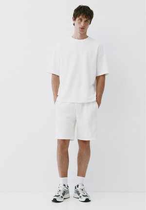 Jeune homme debout les mains dans les poches, portant un t-shirt blanc, un short blanc, des chaussettes blanches et des baskets blanches et noires sur fond uni.