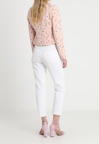 Femme portant un blazer rose à motifs floraux, un jean blanc raccourci avec des ourlets effilochés et des sandales à talons hauts roses, debout devant un fond uni.