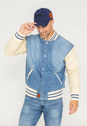 Veste varsity en denim avec des manches crème, des poignets et un col côtelés rayés. Comprend des poches avant et des boutons-pression en argent. Associée à une casquette bleu marine.