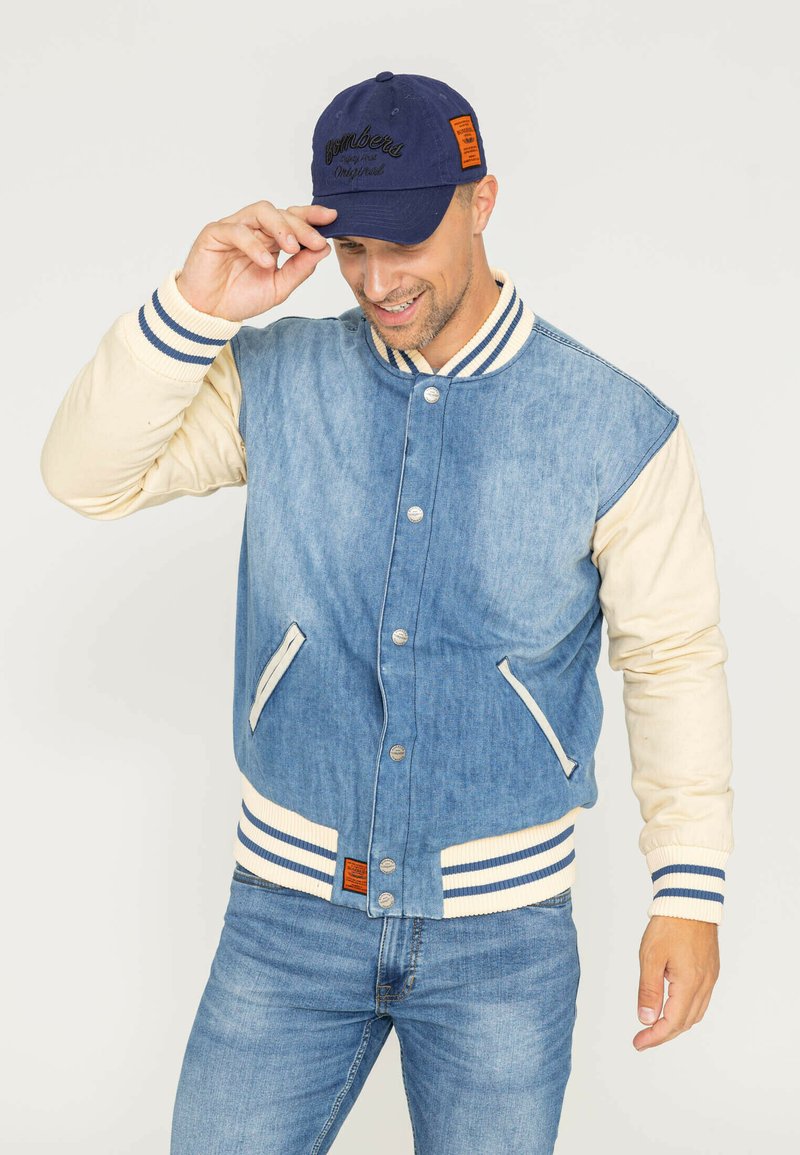 Veste varsity en denim avec des manches crème, des poignets et un col côtelés rayés. Comprend des poches avant et des boutons-pression en argent. Associée à une casquette bleu marine.
