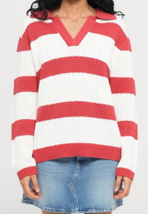 Kobieta ubrana w szeroki, czerwono-biały sweter w paski z dekoltem w kształcie litery V oraz jasnoniebieską spódnicę dżinsową.