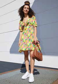 Femme portant une robe fleurie colorée, des baskets blanches et un chouchou, tenant un sac à main marron, debout contre un mur blanc sous la lumière du soleil.