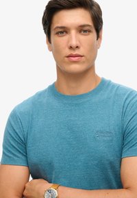 Blauw shirt met korte mouwen van zachte stof, met een klein geborduurd logo op de borst. Het model draagt een gouden polshorloge.