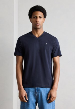 Άνδρας με κοντό αφροαμερικάνικο χτένισμα, που φοράει μπλε ναυτικό T-shirt με V-λαιμόκοψη και μικρό λευκό λογότυπο, καθώς και ανοιχτόχρωμα τζιν, στέκεται μπροστά σε απλό φωτεινό φόντο.