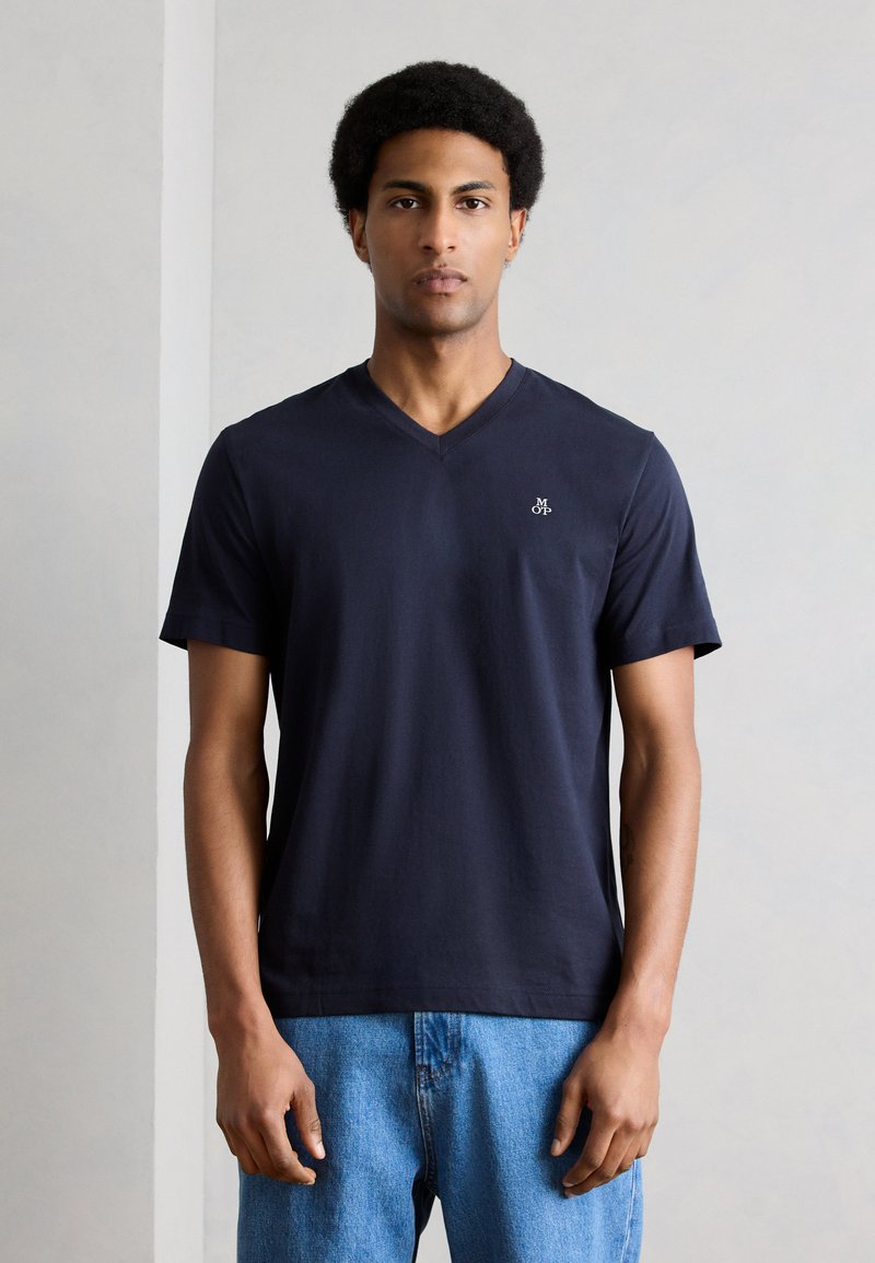 Άνδρας με κοντό αφροαμερικάνικο χτένισμα, που φοράει μπλε ναυτικό T-shirt με V-λαιμόκοψη και μικρό λευκό λογότυπο, καθώς και ανοιχτόχρωμα τζιν, στέκεται μπροστά σε απλό φωτεινό φόντο.