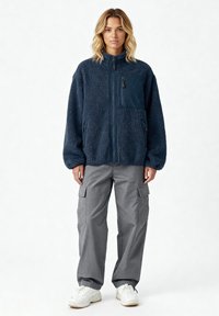 Veste polaire bleu marine avec fermeture éclair devant, dotée d'une poche en tissu lisse contrastant et de deux poches latérales, portée avec un pantalon cargo gris.
