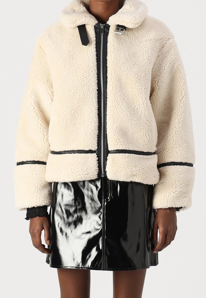 Veste en sherpa polaire couleur crème avec bordure noire portée sur une jupe en vinyle noir brillant, montrée sur une personne des épaules jusqu'au milieu des cuisses.