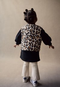 Niño con chaleco acolchado de estampado de leopardo sobre una camiseta negra, pantalones acampanados de pana beige y zapatillas grises, con el cabello peinado en varios nudos.