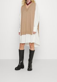 Gilet en tricot beige à col en V sur une robe chemise blanche, associé à des bottes noires jusqu'aux genoux avec une semelle épaisse et un détail de fermeture éclair sur le côté.
