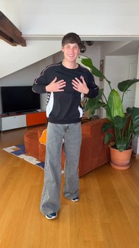 Tânăr zâmbitor, îmbrăcat lejer, gesticulează cu mâinile pe piept într-un living modern cu plante și podea din lemn.