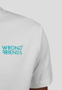 Bílý bavlněný tričko s aqua modrým logem "WRONG FRIENDS" na levé straně hrudi, s krátkým rukávem a klasickým kulatým výstřihem.