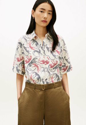 Kvinde iført en kortærmet skjorte med knapper i pink og sort leopardmønster, stukket ned i højtaljede brune bukser med bæltespænde.