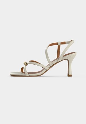 Sandale à talon haut beige avec des fines lanières croisées sur le pied et une petite boule décorative dorée près des orteils.