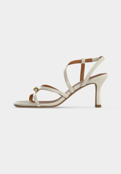 Sandale à talon haut beige avec des fines lanières croisées sur le pied et une petite boule décorative dorée près des orteils.