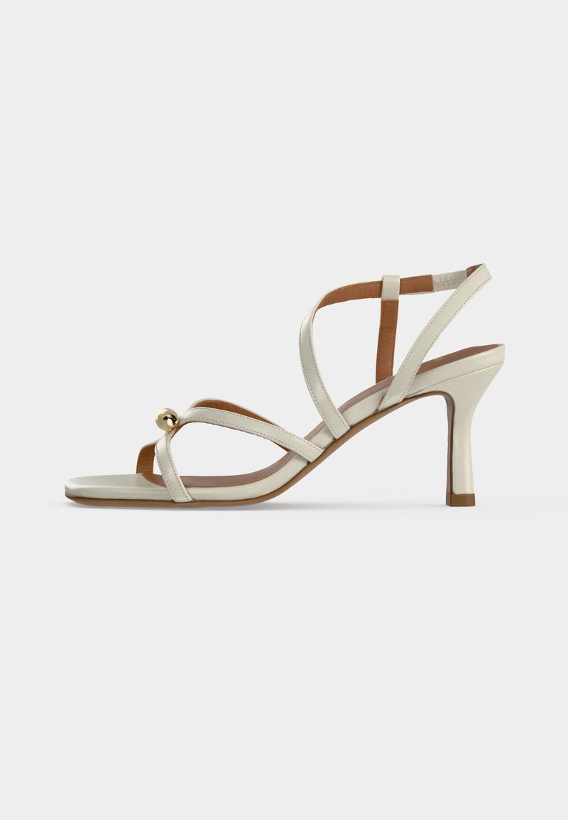 Sandale à talon haut beige avec des fines lanières croisées sur le pied et une petite boule décorative dorée près des orteils.