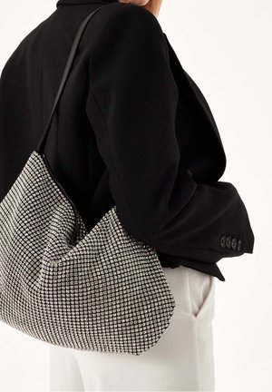 Sølvmesh håndtaske med sorte detaljer, der har et samlet design og en justerbar rem, båret over en sort blazer.