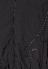 Fekete, könnyű súlyú dzseki középen cipzárral, ívelt varrással, fényvisszaverő csíkkal és kis hímzett logóval a jobb oldalon.