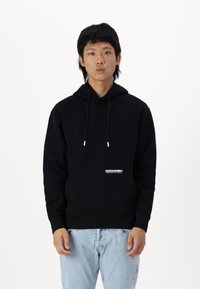 Sweat-shirt à capuche noire avec une poche avant, logo blanc en bas à gauche, cordons de serrage ajustables et poignets en côte. Porté avec un jean bleu clair.