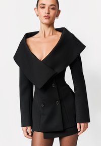 Czarna sukienka w formie marynarki na miarę, z szerokim, drapowanym kołnierzem, dwurzędowymi guzikami, fakturowaną tkaniną, dopasowaną talią i zbudowanymi ramionami.