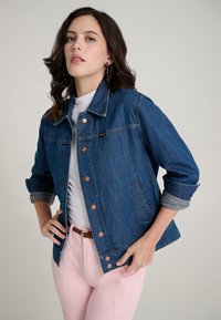 Tamsiai mėlynas džinsinis švarkas su apykakle, variniais mygtukais ir užlenktomis rankovėmis. Dėvimas ant balto viršaus ir derinamas su rožinėmis kelnėmis.