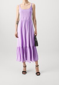 Vestido maxi morado con un cuerpo fruncido, falda acampanada y dobladillo con volantes. Combinado con un bolso negro texturizado y tacones negros.