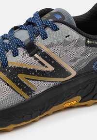 Grå terrengløpesko med teksturert overdel, svarte detaljer og blå lisser. Har gulllogo og Vibram såle for grep.