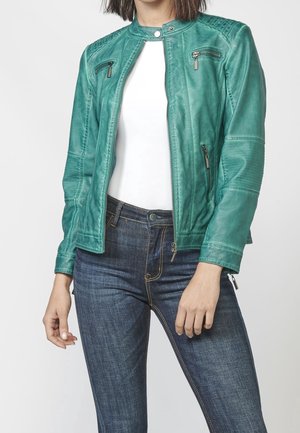 Femme portant une veste en cuir bleu sarcelle par-dessus une chemise blanche et un jean bleu foncé, debout avec une main détendue et l’autre légèrement pliée.
