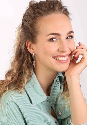 Silberne Anhängerohrringe mit Kettenakzenten und kleinen Herzdetails. Das Modell trägt eine mintgrüne Cordjacke. Lockige Haare umspielen das Gesicht.
