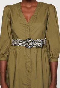 Wpleciony czarny i kremowy pasek z okrągłą czarną klamrą, noszony na oliwkowej sukience z guzikami, z bufiastymi rękawami i dekoltem w serek.