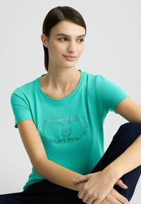Turquoise t-shirt met korte mouwen en een ronde halslijn, versierd met een glanzend grafic logo op de voorkant. Het materiaal lijkt glad en licht van gewicht.