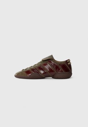 Zapato verde oliva con acentos burdeos brillantes y suela de goma texturizada; presenta un distintivo diseño de tres franjas y cordones en la parte delantera.